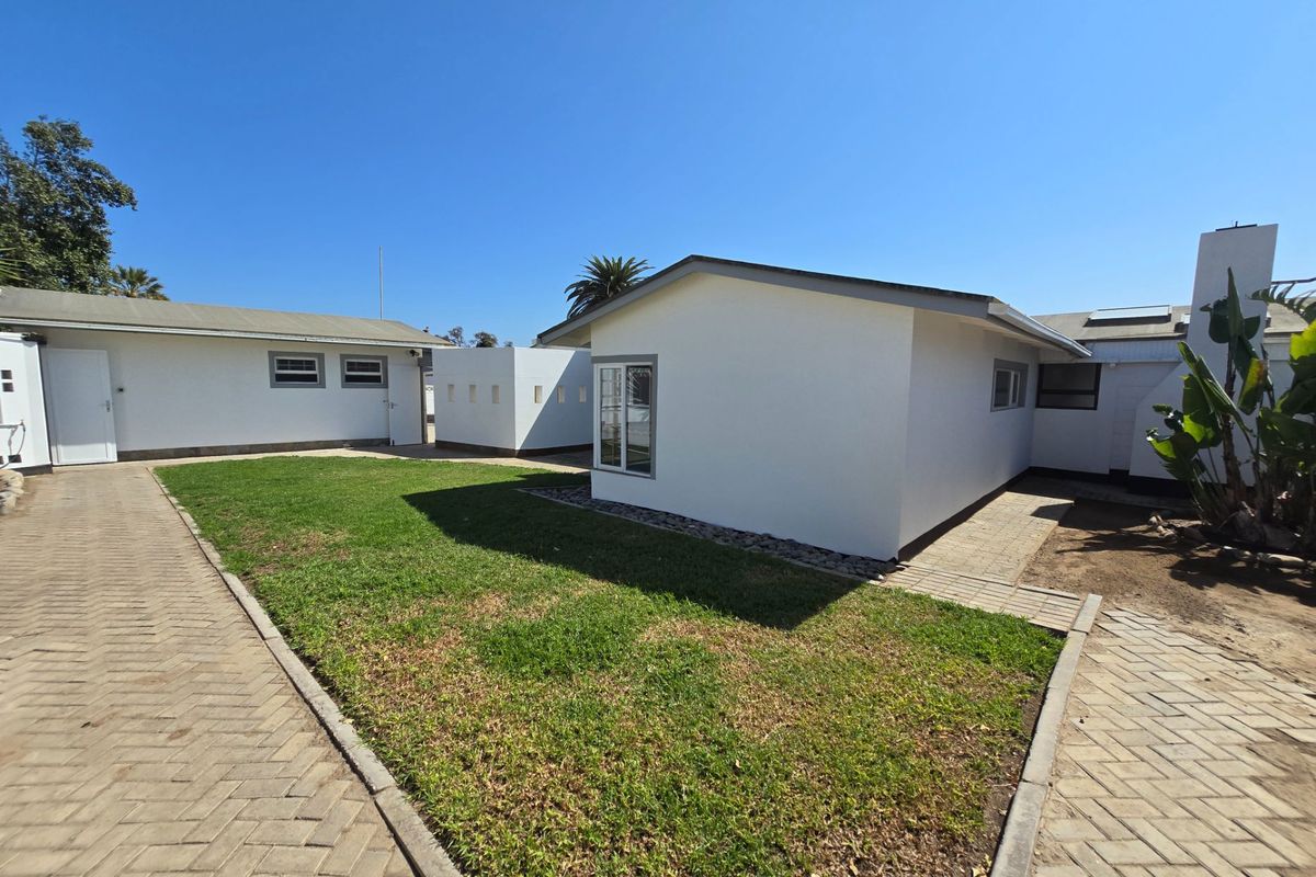 Rear view of the property featuring a well-maintained lawn, paved pathways, and clean white exterior walls under a clear blue sky.
