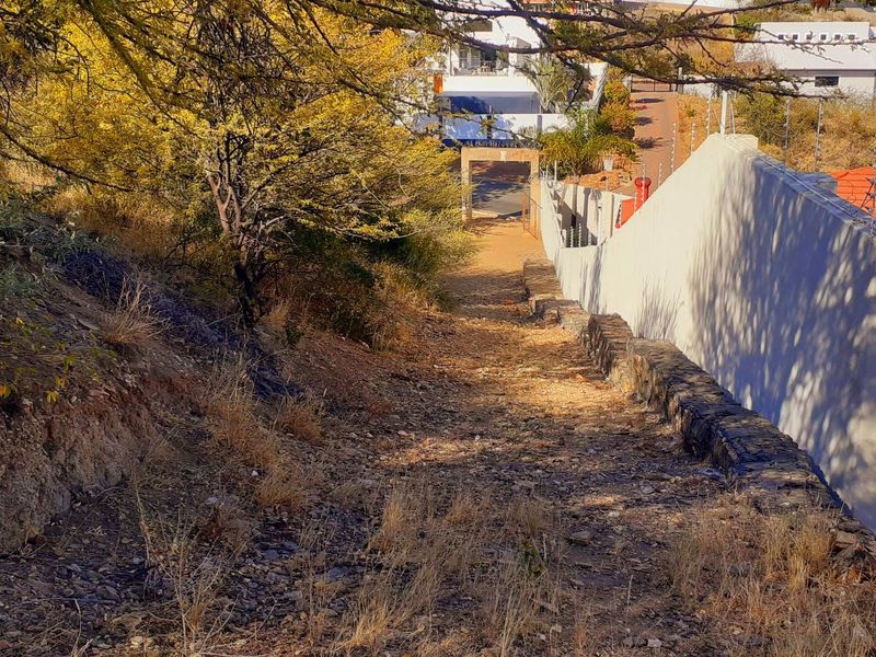 A sloped, undeveloped residential plot featuring a stone retaining wall, electric fencing, and natural vegetation, bordering an existing residential property.