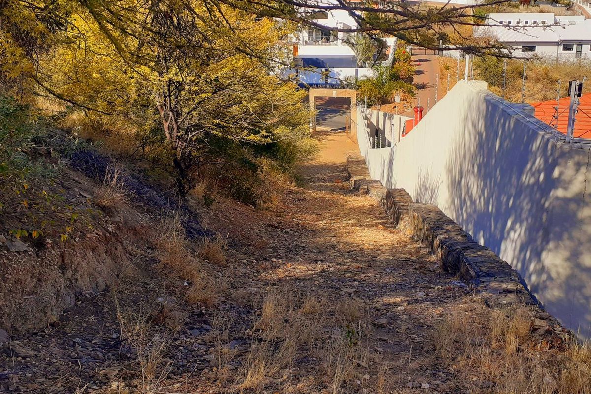 A sloped, undeveloped residential plot featuring a stone retaining wall, electric fencing, and natural vegetation, bordering an existing residential property.