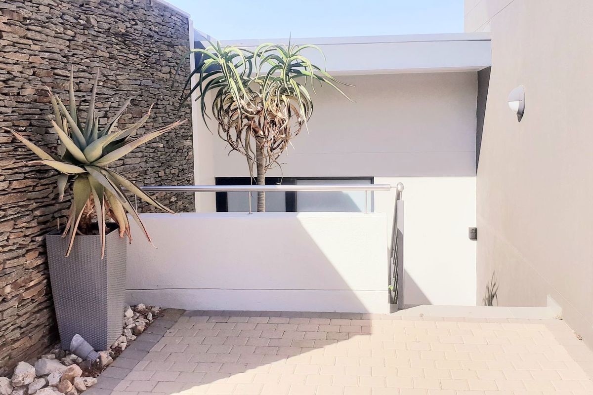 A modern outdoor patio area featuring a textured stone feature wall, paved flooring, and a sleek stainless steel balustrade.