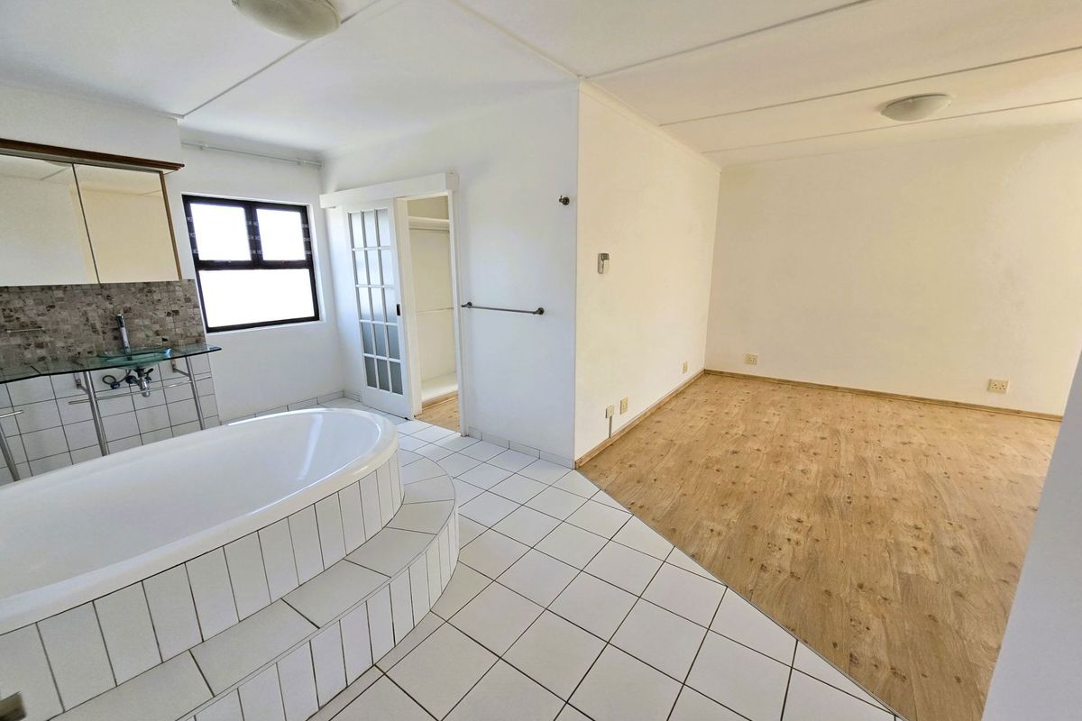Open-plan bedroom and bathroom suite featuring a raised tiled bathtub, glass vanity, and integrated closet space with wood-look flooring.