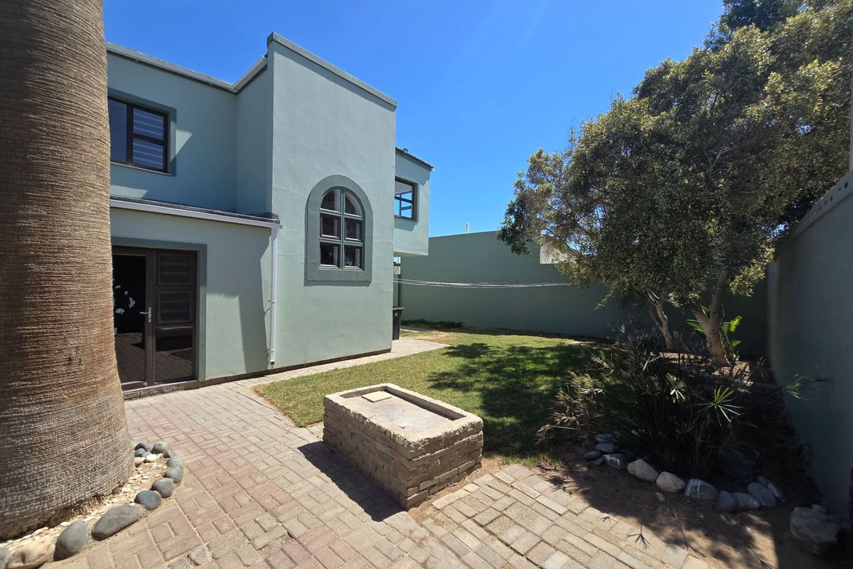 A private rear garden featuring a paved patio area, built-in brick braai, and established lawn, accessible via double doors from the main house.