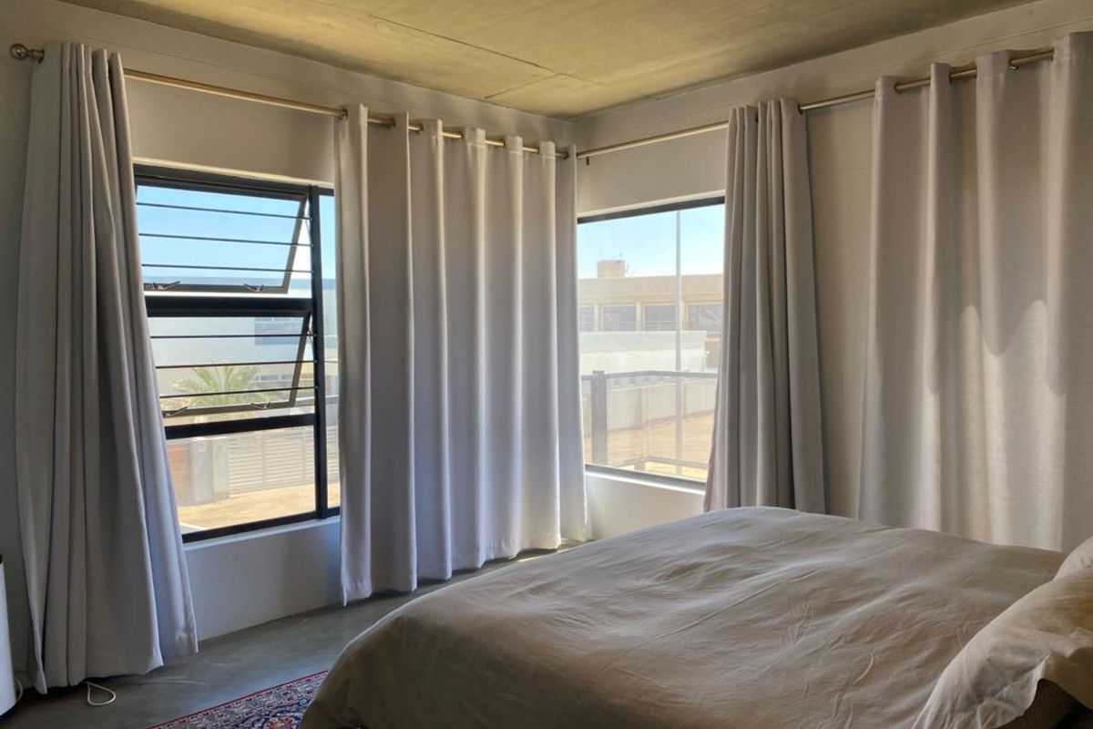 A bright bedroom featuring large windows with security bars, neutral curtains, and a concrete ceiling, offering a clean and modern aesthetic.