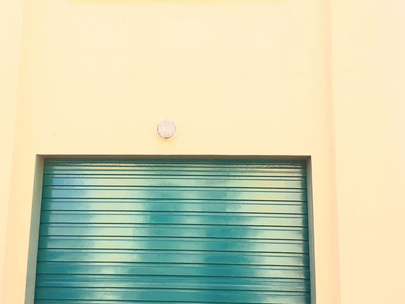 Exterior view of a garage featuring a green roller shutter door, an exterior wall light, and a grid-patterned window above.