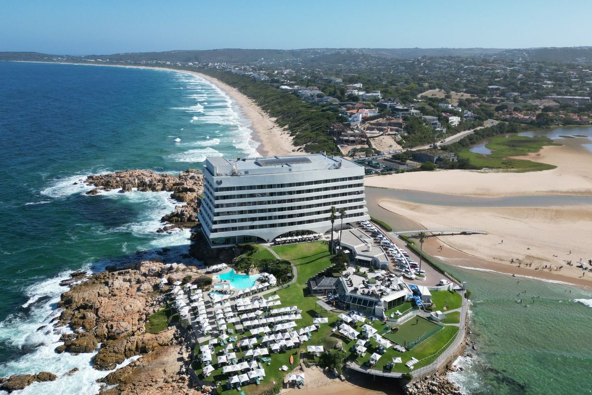 Aerial view of a coastal property featuring a large multi-story building, swimming pool, landscaped grounds, and direct access to the beach and ocean.