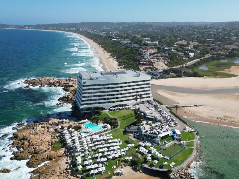 Aerial view of a coastal property featuring a large multi-story building, swimming pool, landscaped grounds, and direct access to the beach and ocean.