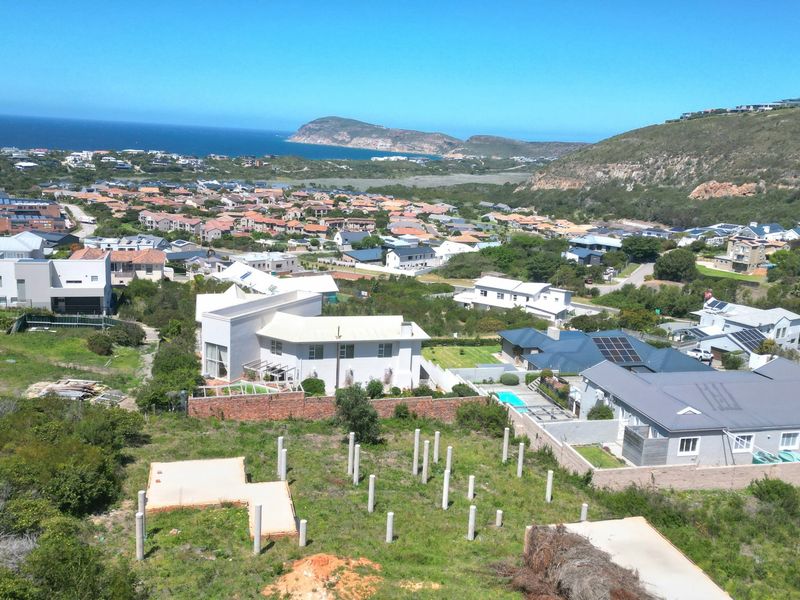 Aerial view of a residential estate featuring a vacant plot with prepared concrete foundation pillars and a slab, overlooking the surrounding coastal neighborhood.