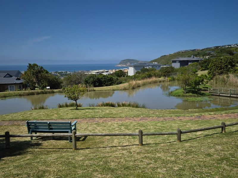 A scenic view of a landscaped pond with a wooden footbridge, surrounded by manicured lawns and coastal hillside vistas.