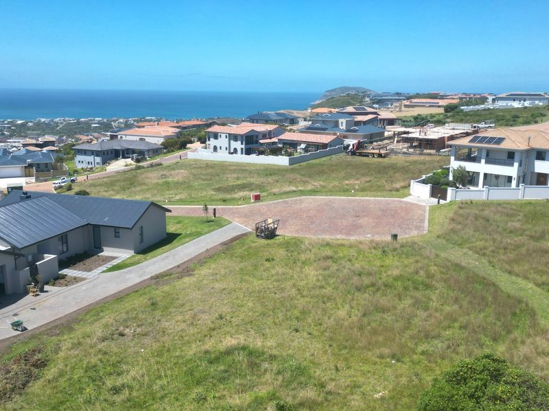 An elevated aerial view of a residential vacant land plot featuring a paved driveway, overlooking a coastal neighborhood and the ocean.