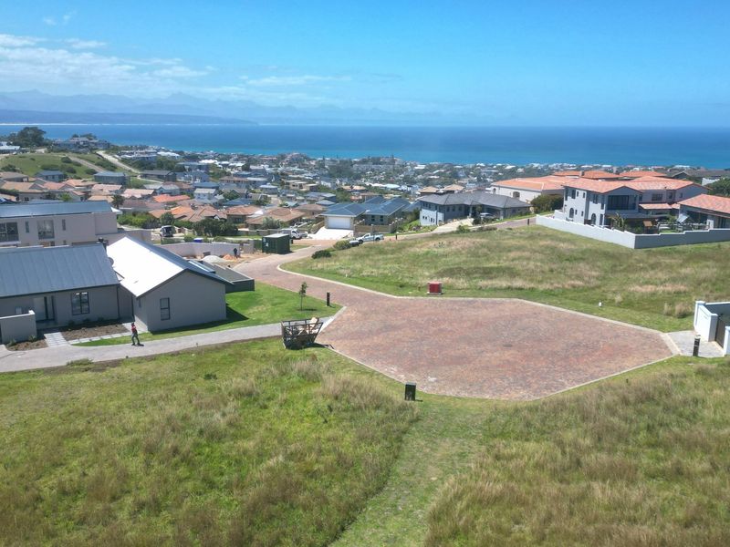 Aerial view of a vacant residential plot featuring a paved driveway, situated in a coastal estate with panoramic ocean and mountain views.