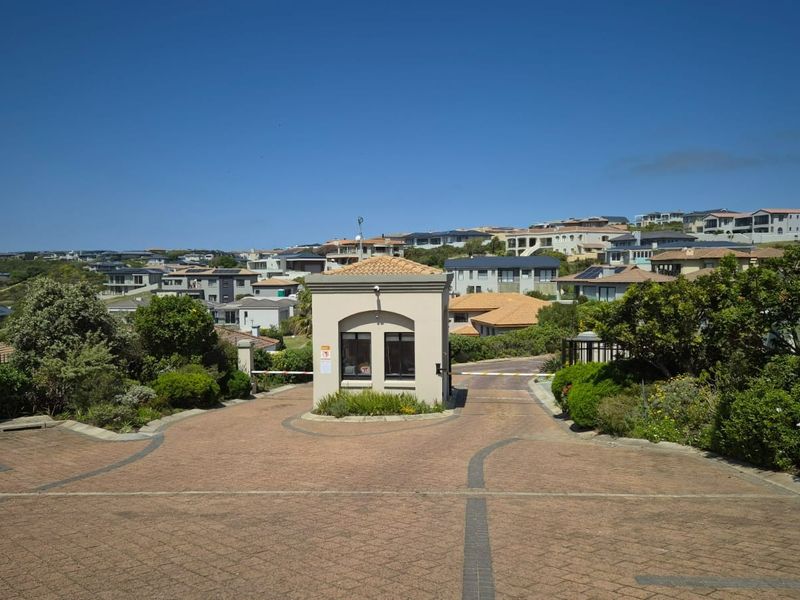 A well-maintained residential estate entrance featuring a security gatehouse, paved driveway, and established landscaping within a secure community setting.