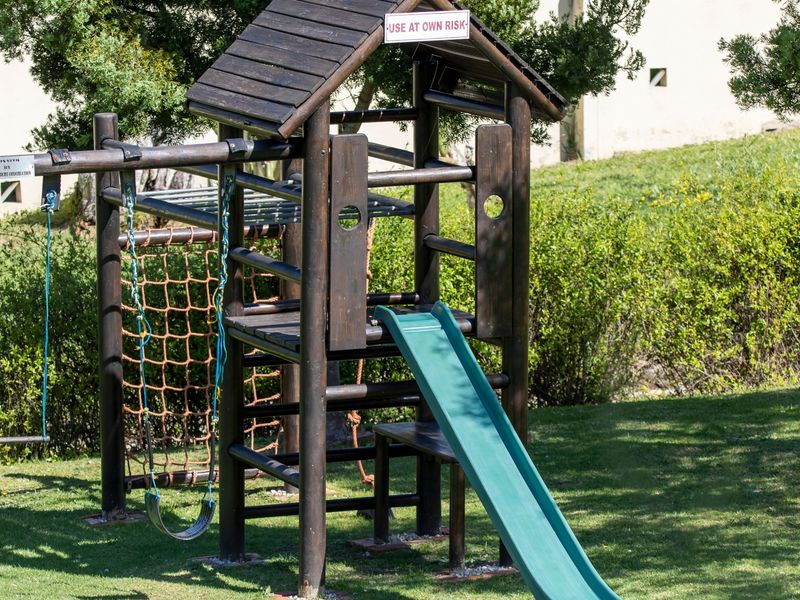 A residential garden area featuring a wooden jungle gym with a slide, swing set, and climbing net, set within a landscaped lawn.