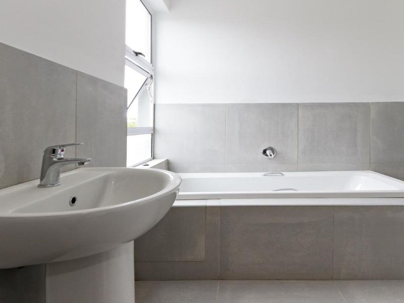 A clean, modern bathroom featuring a white pedestal basin, a built-in bathtub with grey tiled surround, and neutral wall finishes.