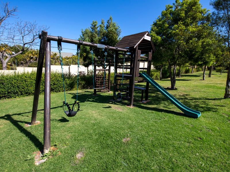 A well-maintained residential garden featuring a wooden playground structure with a slide, swings, and climbing net set within a spacious lawn area.