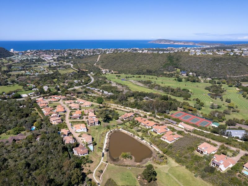 Aerial view of a residential estate featuring a golf course, tennis courts, and a central lake with coastal views in the background.
