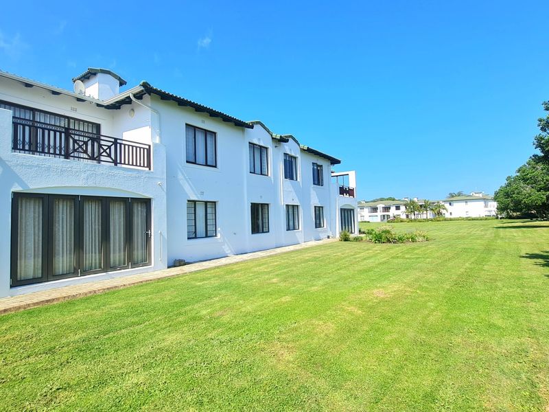 Rear view of a multi-story apartment building featuring white exterior walls, a tiled roof, balconies, and extensive well-maintained lawn areas.