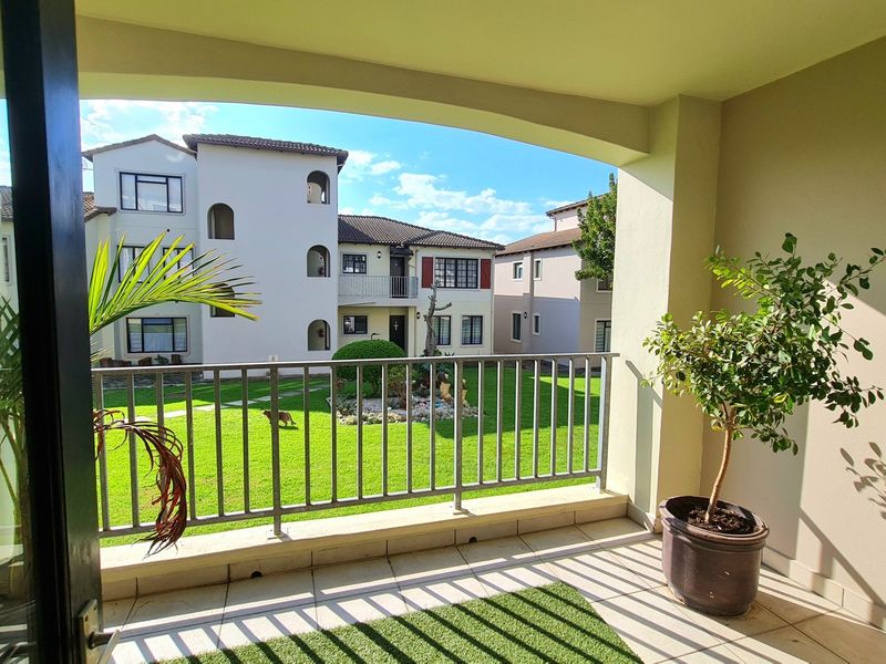 A covered balcony featuring tiled flooring and a metal balustrade, offering a pleasant view over the complex's communal garden area.