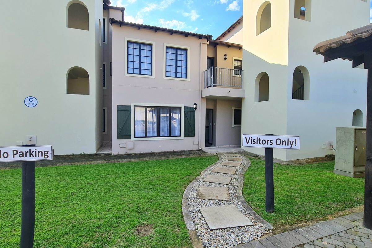 A view of the apartment building exterior featuring a stone paver pathway leading to the entrance, a small balcony, and landscaped garden area.