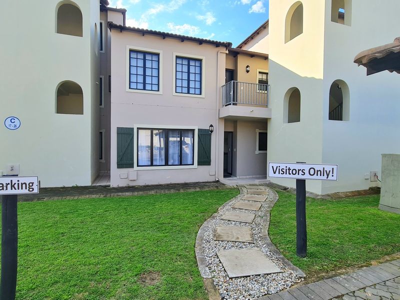 A view of the apartment building exterior featuring a stone paver pathway leading to the entrance, a small balcony, and landscaped garden area.