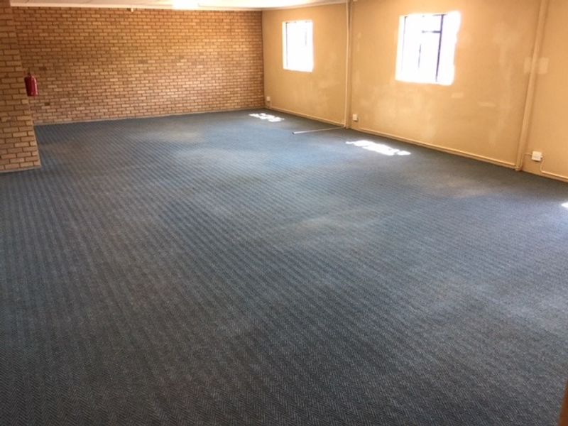A spacious, empty office area featuring exposed brickwork, carpeted flooring, and natural light from multiple windows.