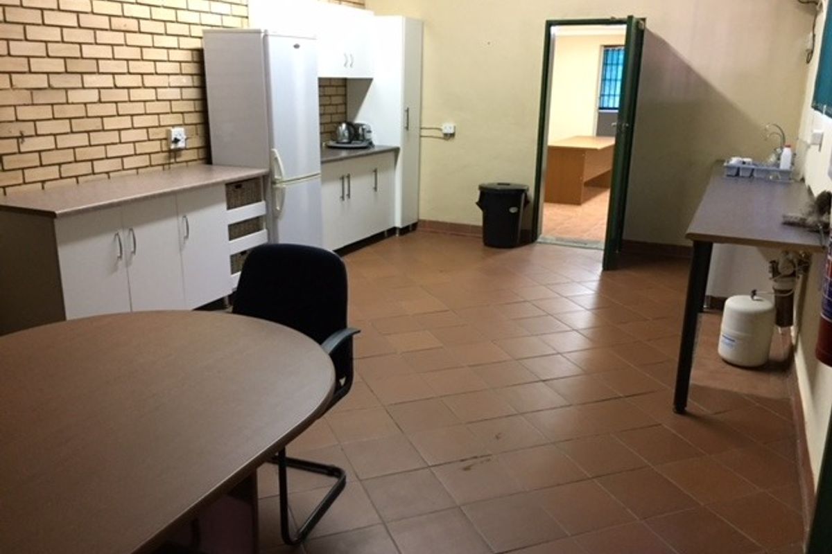 A functional office kitchenette featuring built-in cabinetry, laminate countertops, tiled flooring, and an exposed brick feature wall.