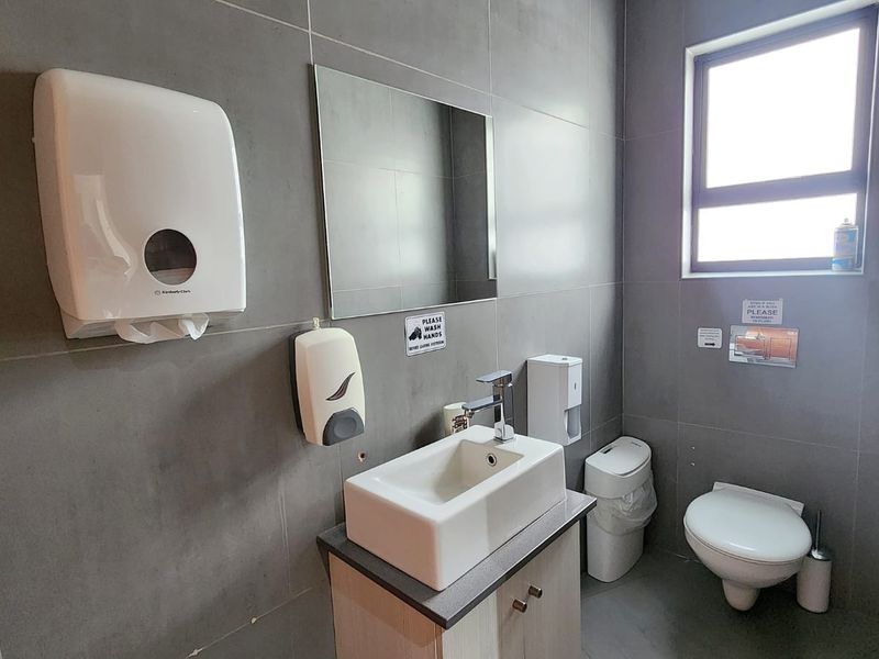 A functional office bathroom featuring grey tiled walls, a modern wall-mounted basin with vanity cabinet, and a standard toilet.