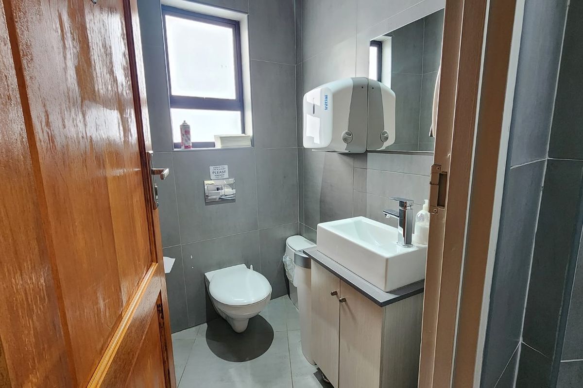 A modern, compact office bathroom featuring grey wall tiles, a vanity cabinet with a vessel sink, and a wall-mounted toilet.