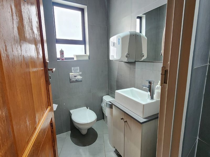 A modern, compact office bathroom featuring grey wall tiles, a vanity cabinet with a vessel sink, and a wall-mounted toilet.