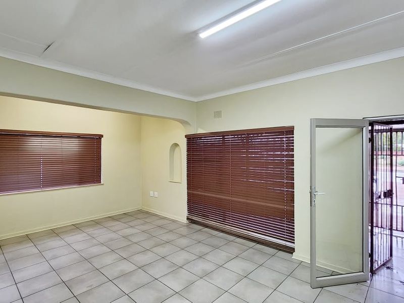 A vacant office space featuring tiled flooring, large windows with venetian blinds, and a secure entrance door with a metal security gate.