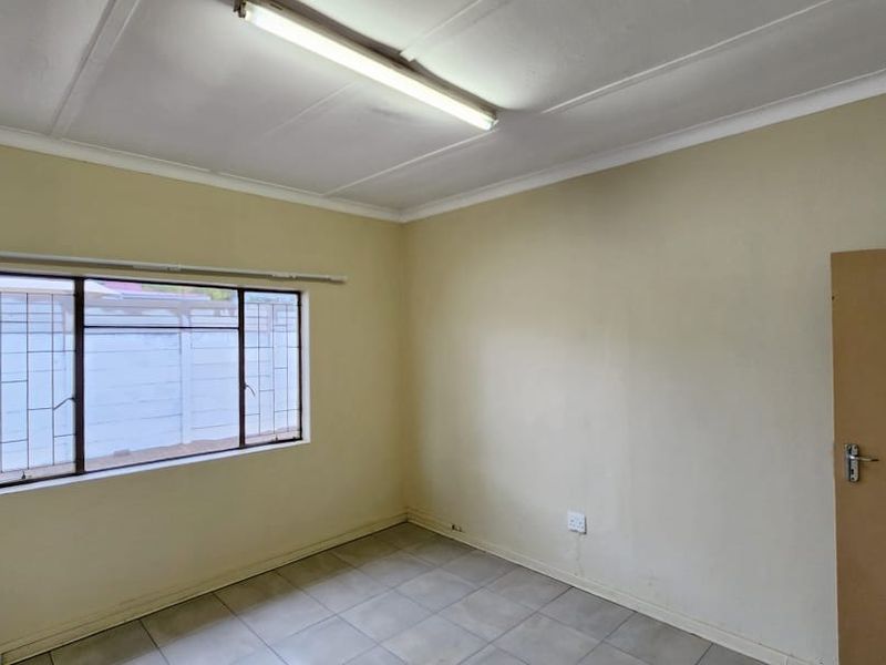 A vacant office space featuring tiled flooring, a fluorescent ceiling light, and a window fitted with security bars.