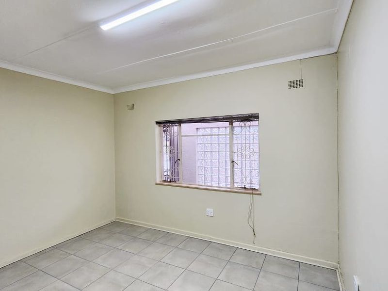 A clean, empty office space featuring tiled flooring, a fluorescent light fixture, and a window with security bars.
