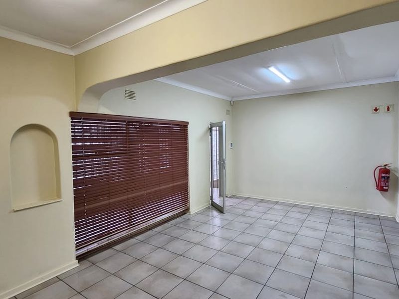 A bright, open-plan office space featuring tiled flooring, a decorative wall niche, and large windows with venetian blinds.