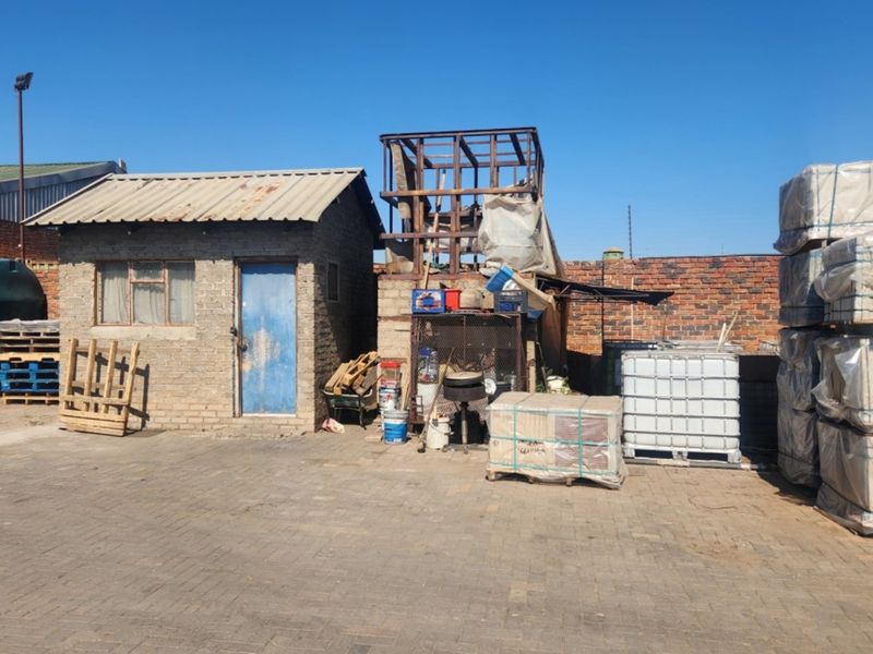 Industrial yard area featuring a small brick utility structure, paved ground, perimeter walling, and various storage tanks and industrial materials.