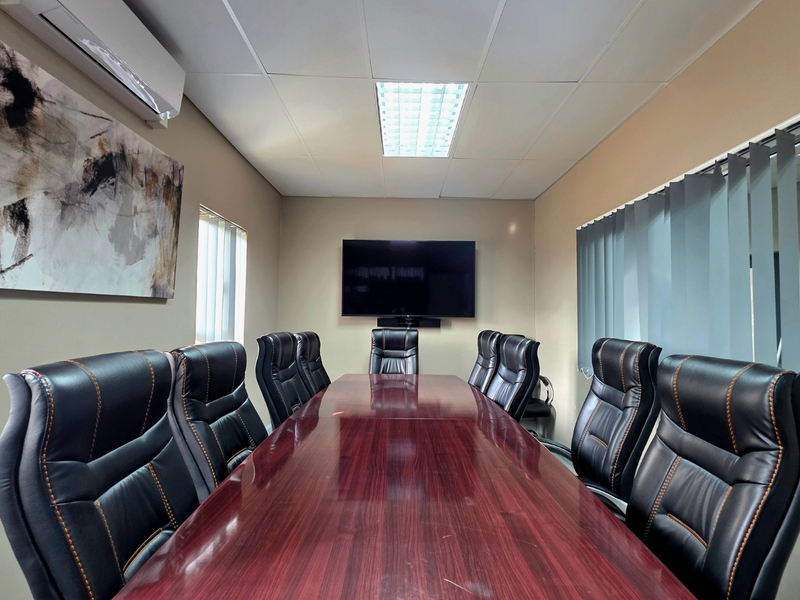 A professional boardroom featuring a large meeting table, wall-mounted display screen, suspended ceiling with recessed lighting, and climate control.