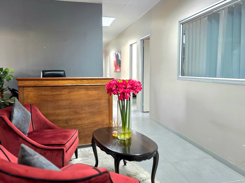 A professional office reception area featuring a wooden reception desk, tiled flooring, and a bright, clean interior layout with access to adjacent office spaces.