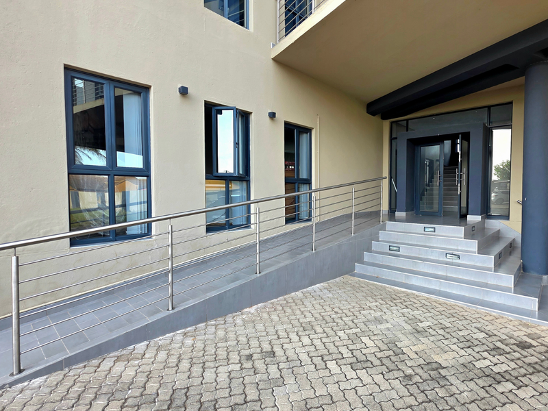 Modern office building entrance featuring a tiled wheelchair ramp, stainless steel handrails, and illuminated concrete steps leading to a glass-fronted doorway.