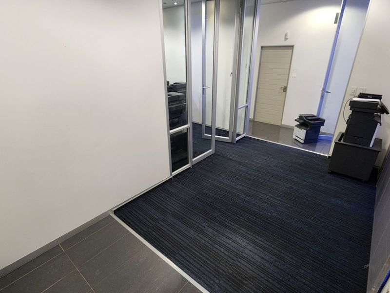 A modern office space featuring glass partitions, a mix of carpet and tiled flooring, and recessed lighting, providing a clean and professional work environment.