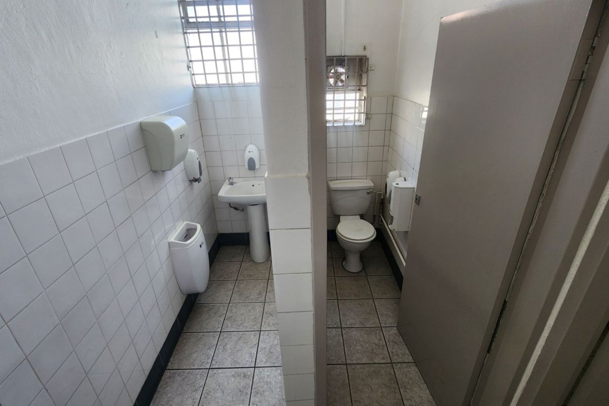 A functional commercial bathroom facility featuring a pedestal sink and a separate toilet cubicle with tiled finishes and security bars on the windows.