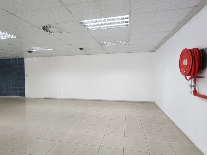 An empty retail or commercial space featuring ceramic floor tiles, a suspended ceiling with recessed lighting, and a wall-mounted fire hose reel.