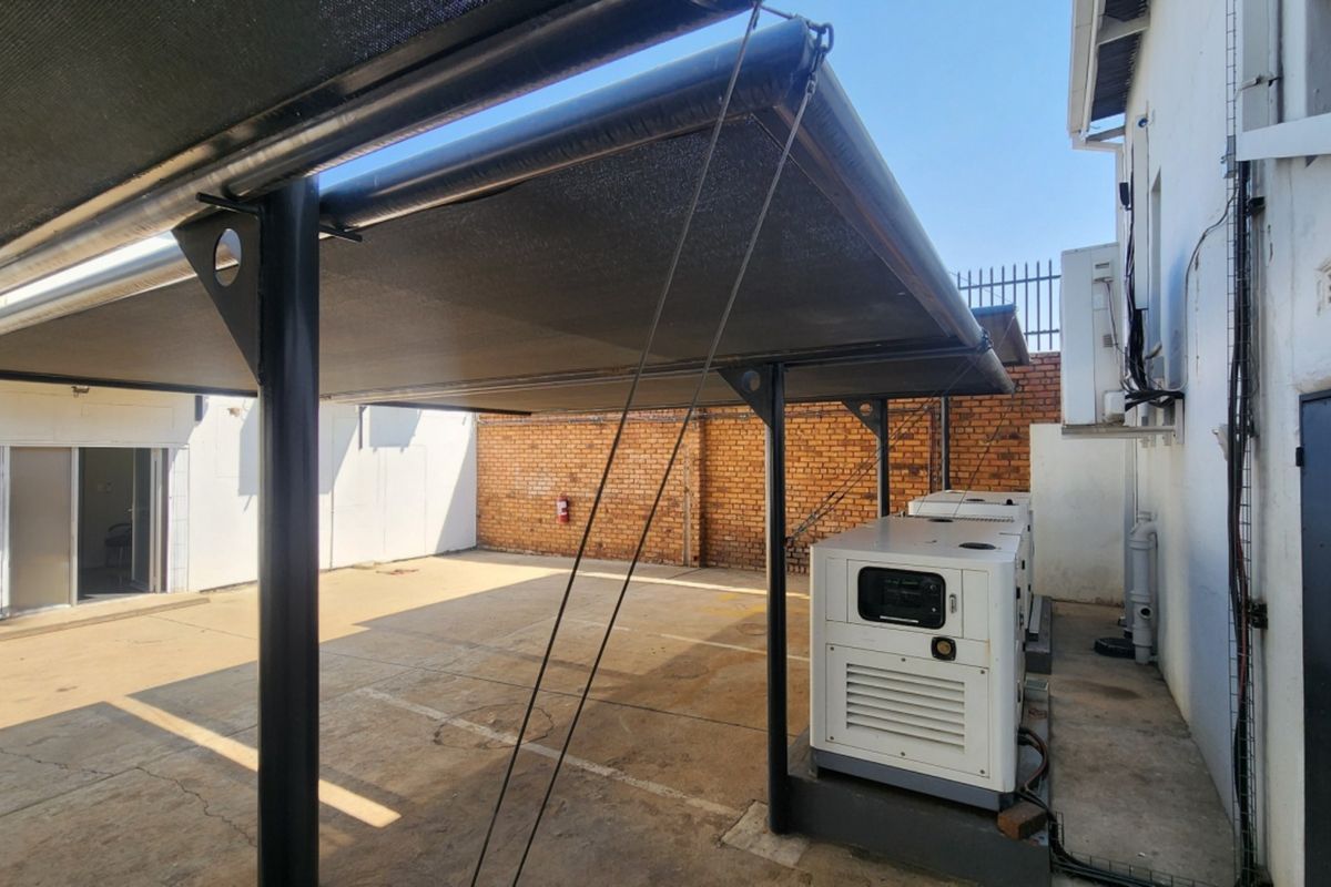 Rear exterior area featuring a shade cloth carport, concrete paving, and a backup generator unit against a brick boundary wall.