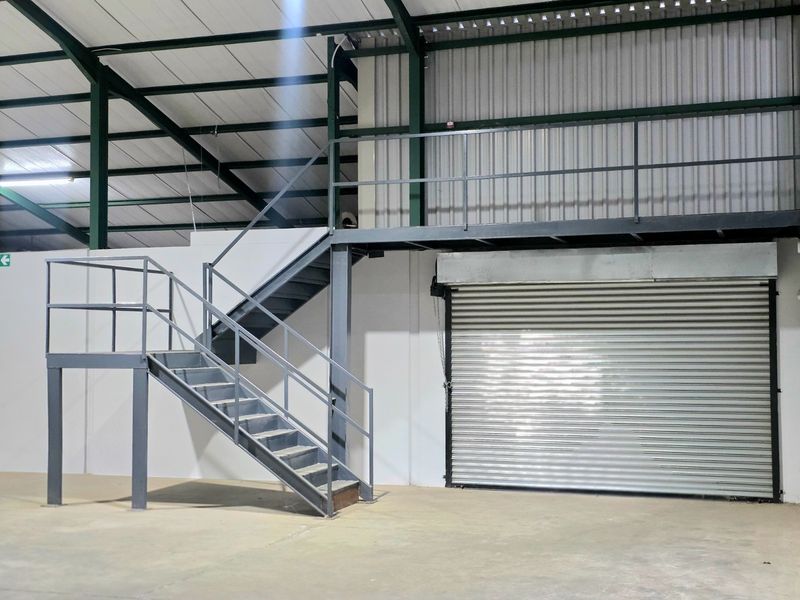 Industrial warehouse space featuring a steel mezzanine level, staircase, and a large roller shutter door with high ceilings and concrete flooring.