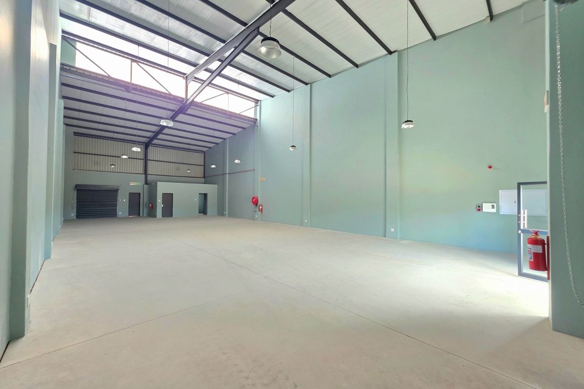 Spacious industrial warehouse unit featuring high ceilings with exposed steel trusses, concrete flooring, natural light via skylights, and a roller shutter door.