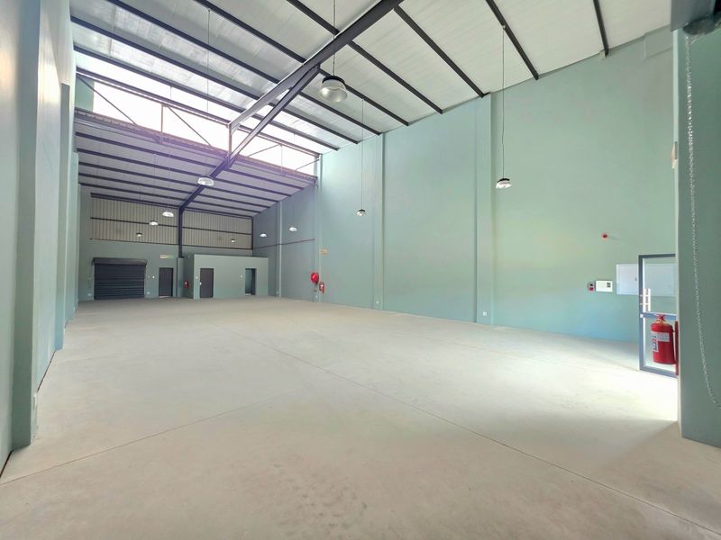 Spacious industrial warehouse unit featuring high ceilings with exposed steel trusses, concrete flooring, natural light via skylights, and a roller shutter door.