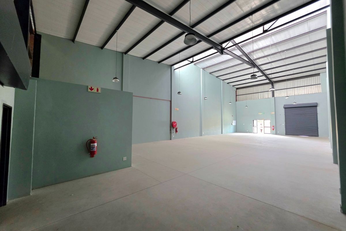 Spacious industrial warehouse unit featuring high ceilings with exposed steel trusses, concrete flooring, roller shutter access, and natural light through translucent roof panels.