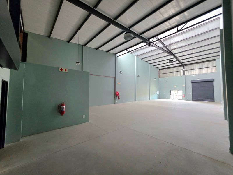 Spacious industrial warehouse unit featuring high ceilings with exposed steel trusses, concrete flooring, roller shutter access, and natural light through translucent roof panels.