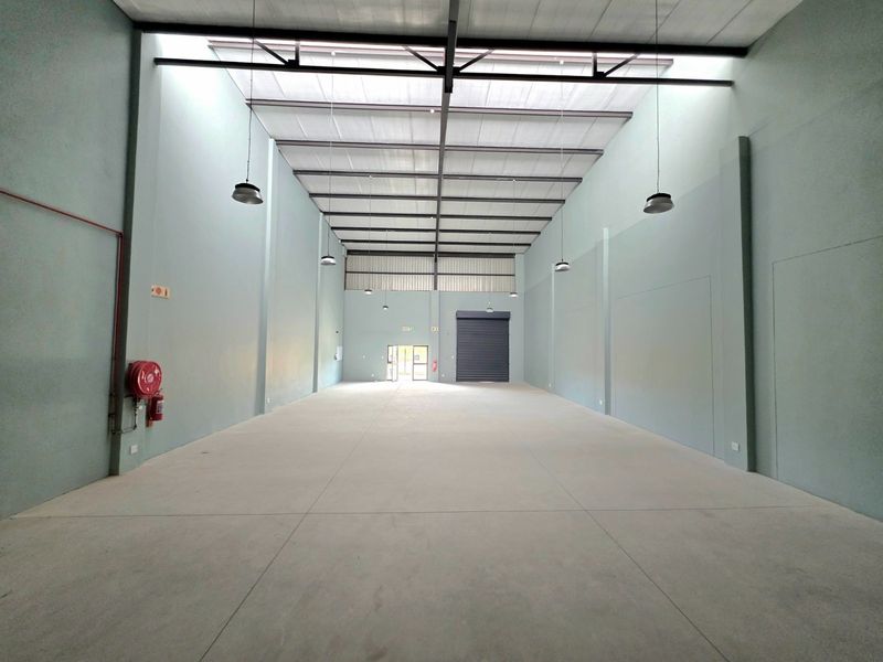 Spacious industrial warehouse unit featuring high ceilings with exposed steel trusses, concrete flooring, roller shutter door access, and integrated fire safety equipment.