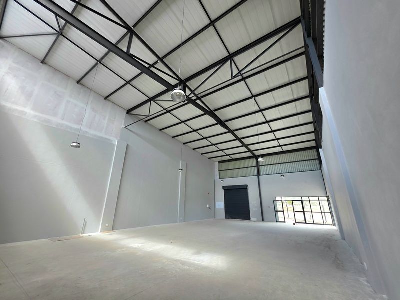 Spacious industrial warehouse unit featuring high ceilings with exposed steel trusses, concrete flooring, and a large roller shutter door for easy access.