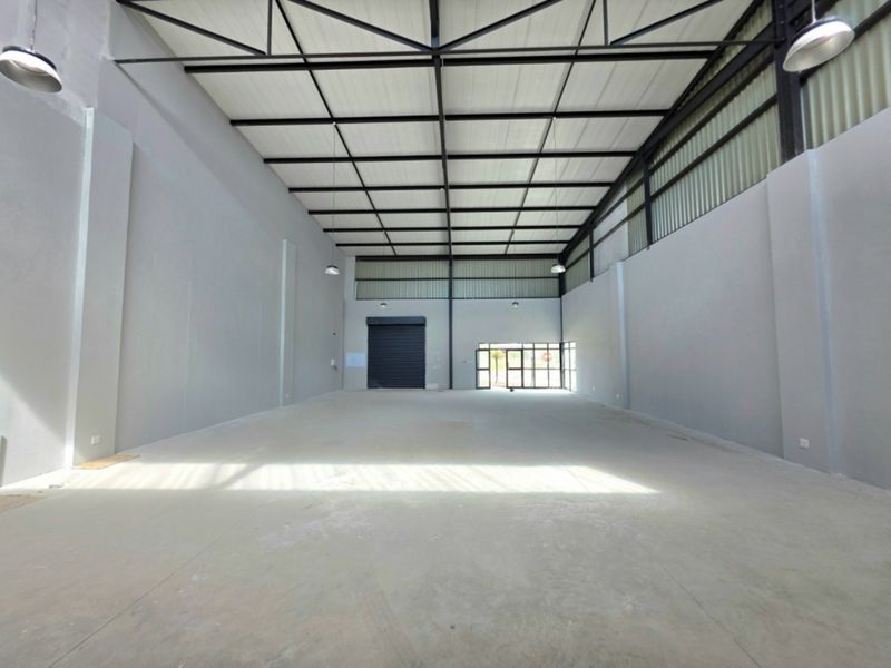 Spacious industrial warehouse unit featuring high ceilings with exposed steel trusses, concrete flooring, and a large roller shutter door for easy access.