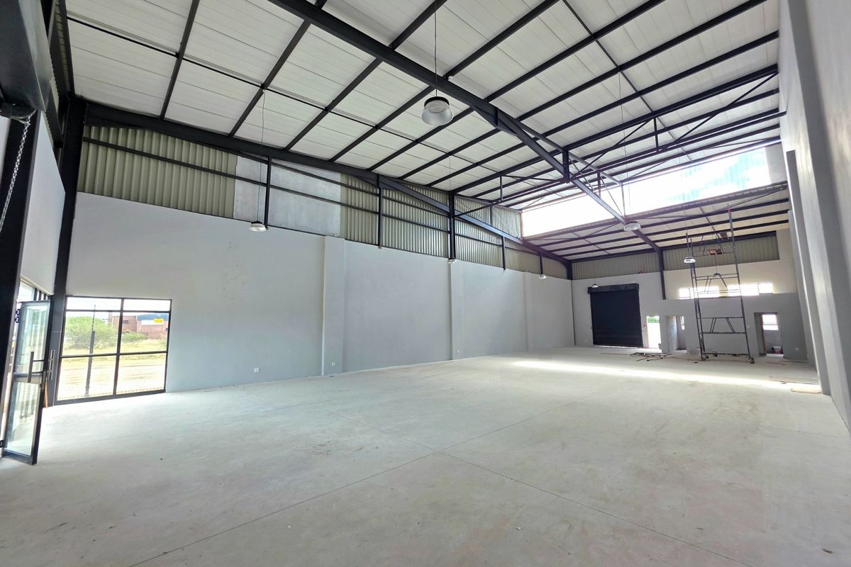 Spacious industrial warehouse unit featuring high ceilings with exposed steel trusses, concrete flooring, and a large roller shutter door for easy access.