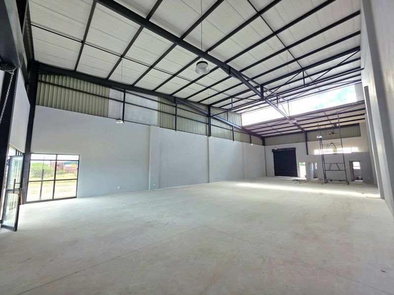 Spacious industrial warehouse unit featuring high ceilings with exposed steel trusses, concrete flooring, and a large roller shutter door for easy access.